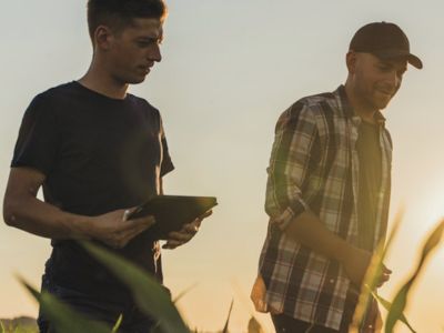 Due persone camminano in un campo coltivato al tramonto: una consulta un tablet e l’altra indossa una camicia a quadri e cappellino