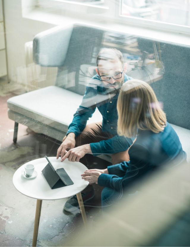 man and woman sitting and discussing business matters. Tablet on desk in front of them. 