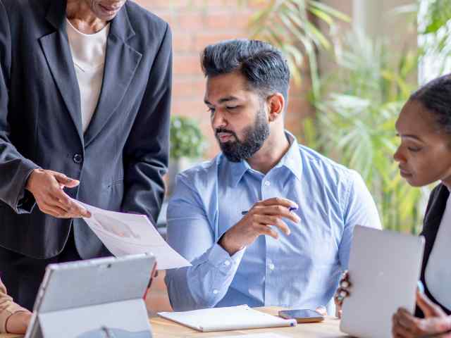 3 employees reviewing work. One elder employee is standing and showcasing a paper to 2 other employees siting at a desk