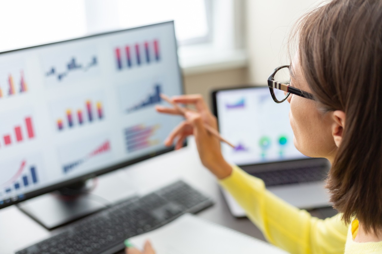 woman flipping through a folder with a pencil in her hand with a monitor in front of her. She seems to be checking through the information in the folder with what is on the monitor.