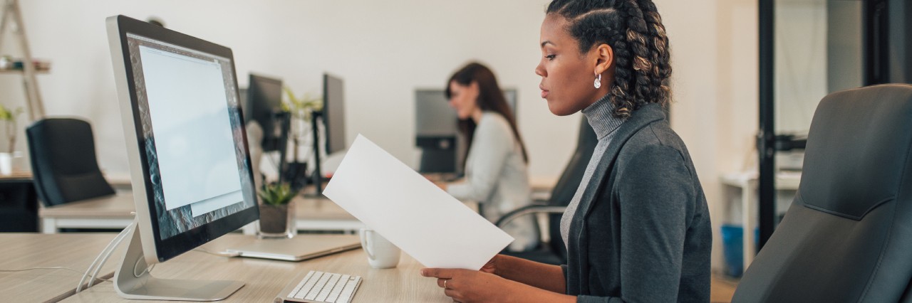 woman flipping through a folder with a pencil in her hand with a monitor in front of her. She seems to be checking through the information in the folder with what is on the monitor.
