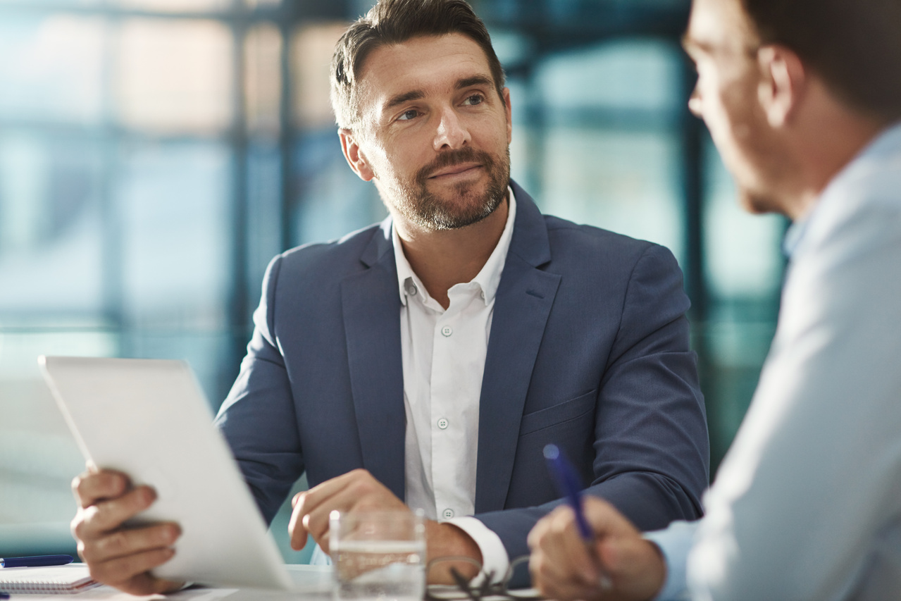 two men discussing something at the table. One is holding a notebook and the other is holding a pen.