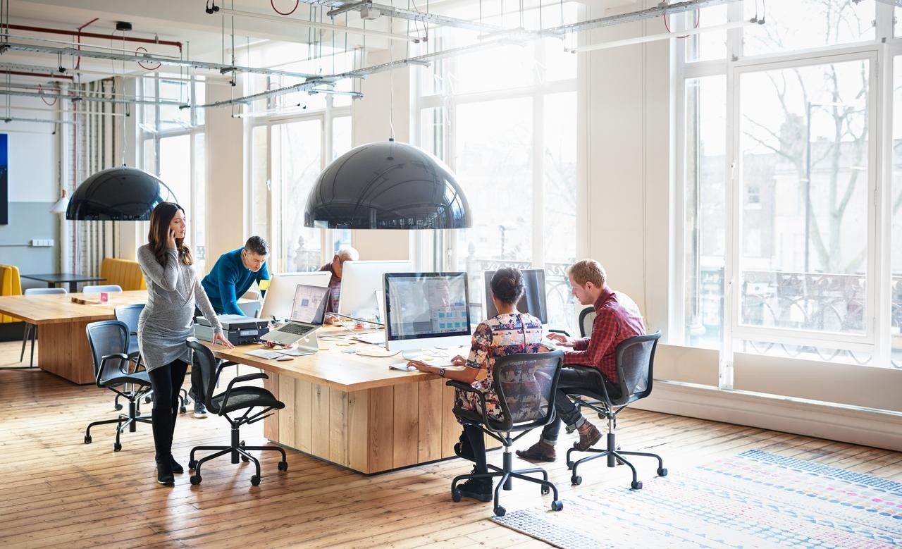 Five people working in a office at a desk together in a well-lit room with large open windows. A man is hidden behind his computer screen. The man furthest away in a blue jacket stands while hunching over his computer screen. A woman in a grey dress is making a phone call standing while looking at her computer. The other two men are facing away working away at their computers.
