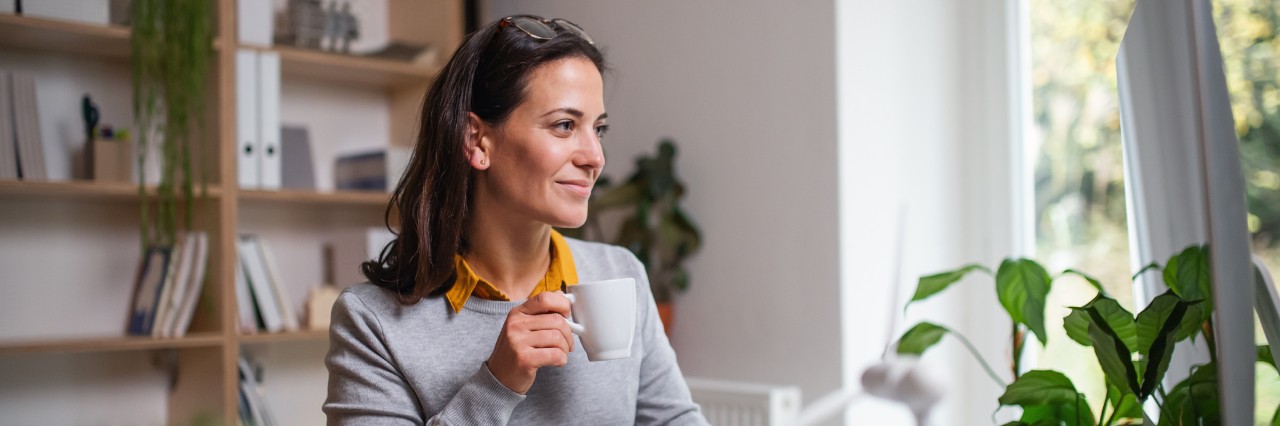 woman in office with plant and bookshelf holding a coffee mug while looking at her monitor