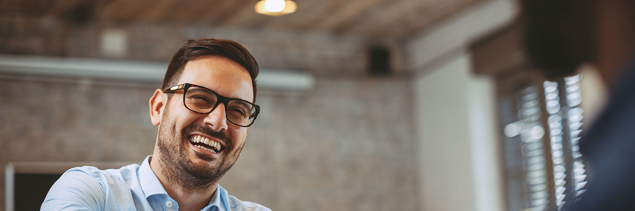 Ein hemdtragender Mann mit Brille lacht herzlich in Richtung seines Gegenübers